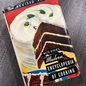 May include: A vintage cookbook titled "Meta Given's Modern Encyclopedia of Cooking" with a new revised edition. The cover features a chocolate cake with white frosting and green sprinkles.
