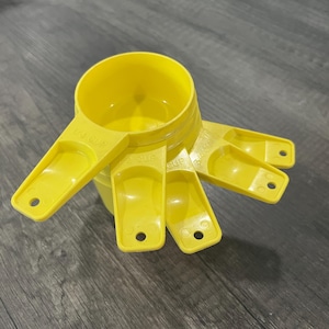 May include: A set of four bright yellow plastic measuring cups stacked together. Each handle features a small circular hole at the end and embossed text indicating the capacity, including 1 cup, 1/2 cup, 1/3 cup, and 1/4 cup measurements.