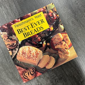 May include: A cookbook titled "Fleischmann's Yeast BEST-EVER BREADS" featuring images of various breads, including a loaf with seeds, sliced bread, croissants, and cinnamon rolls. The cover has a yellow banner with the title.