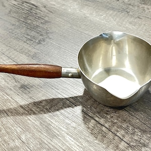 May include: A small, silver-colored saucepan with a wooden handle. The handle is a dark brown color. The saucepan has a spout for easy pouring. The handle is attached to the pan with a metal fitting.