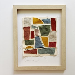 May include: Abstract textile art piece in a light wood frame. The artwork features various fabric patches in shades of red, yellow, green, and blue, stitched onto a cream-colored background. The frame is a natural wood tone.