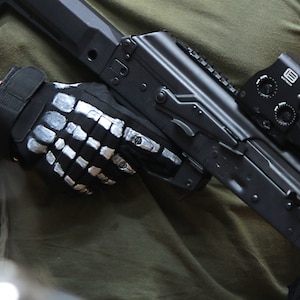 Puede incluir: Un rifle negro con una mira de punto rojo y un guante táctico negro de mano esqueleto. El rifle tiene una culata de polímero negro y un cañón de metal negro.