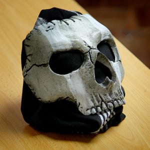 Puede incluir: Una máscara de calavera con una apariencia desgastada y envejecida. La máscara es principalmente blanca y gris, con cuencas de ojos negras y una base de tela negra. El diseño de la calavera presenta grietas y detalles, creando un efecto realista y espeluznante.
