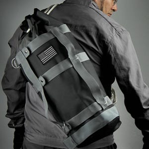 Może przedstawiać: Czarna torba kurierska z płótna z szarymi paskami i małą naszywką z amerykańską flagą. Torba ma zamykanie na rolowanie i wiele klamer.