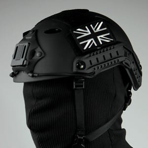 Puede incluir: Un casco táctico negro con un parche de la Union Jack en la parte delantera. El casco tiene un sistema de riel lateral y una correa para la barbilla.