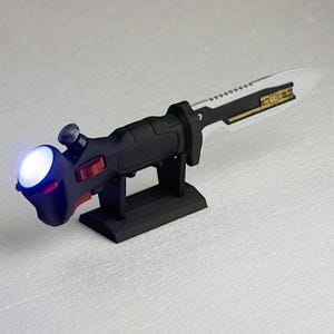 以下が含まれることがあります： 黒と銀のナイフで、透明な刃とハンドルに薄い青色のLEDが付いています。ナイフは黒いスタンドの上に置かれています。