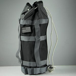 Pode incluir: Uma bolsa de cordão preta com alças cinzas e um remendo da bandeira americana. A bolsa tem um cordão branco e é feita de um material durável.