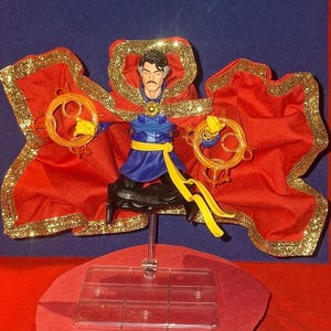Könnte beinhalten: Eine Actionfigur von Doctor Strange, gekleidet in einen roten und goldenen Umhang, mit leuchtenden Ringen. Die Figur ist in der Luft posiert, mit einer dynamischen Haltung.