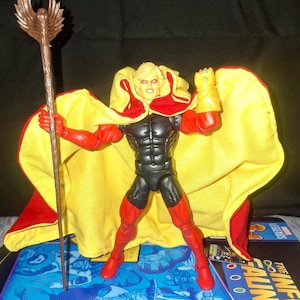 May include: Superhero action figure with a yellow and red cape, black and red suit, and a staff. The figure has blonde hair and is posed with one fist clenched and the other hand holding the staff. The figure is standing on a blue surface.