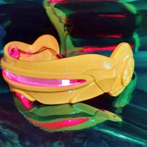 May include: A yellow plastic headband with a pink light-up visor. The headband has a futuristic design and is perfect for a costume or cosplay.