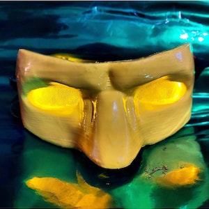 Op de afbeelding: Een geel plastic masker met twee grote, ronde, gele ogen. Het masker heeft een prominente neus en een brede mond.