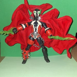May include: Action figure of a character in a black, red, and white costume with a large red cape. The figure has green accents and holds two green, blade-like weapons. The figure is posed against a green background.