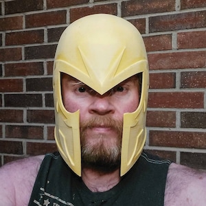 May include: A light yellow helmet with a pointed visor and angular cheek guards. The helmet is worn by a person with a beard and a dark green tank top. The background is a brick wall.