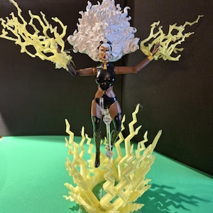 Puede incluir: Figura de acción de Storm con un body negro y plateado, cabello blanco y efectos de rayos amarillos. La figura está posada con los brazos extendidos y está montada sobre un soporte transparente con rayos amarillos.