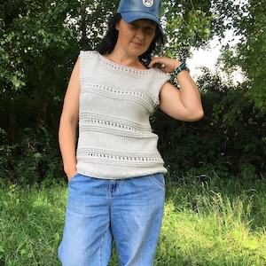 May include: A woman wearing a light gray knitted sleeveless top with a delicate openwork pattern and blue jeans. She is wearing a blue baseball cap with a white logo that says "NYC".