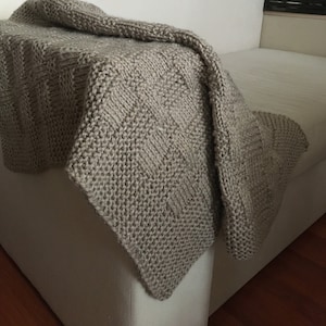 Op de afbeelding: Een beige gebreide deken met een textuurpatroon, gedrapeerd over een witte bank.