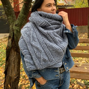 May include: A blue knitted scarf with a textured pattern. The scarf is worn around the neck of a person wearing a denim jacket and jeans.