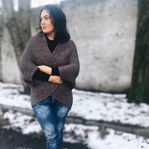 May include: A woman wearing a brown knitted cardigan with a black mock neck shirt and blue jeans. She is standing in a snowy outdoor setting.
