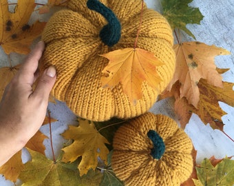 May include: Two knitted pumpkins in a mustard yellow color with green stems. The pumpkins are surrounded by fall leaves in shades of yellow, orange, and brown.