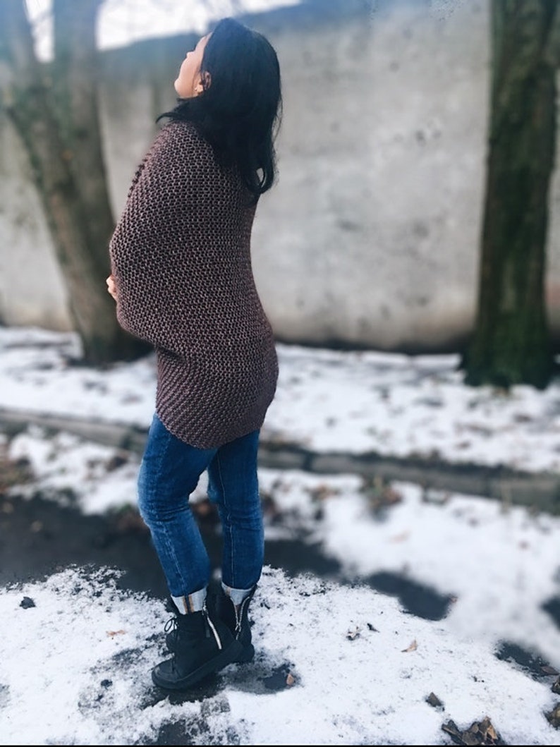 May include: A woman wearing a brown knitted cardigan, blue jeans, and black boots. She is standing in a snowy outdoor setting.