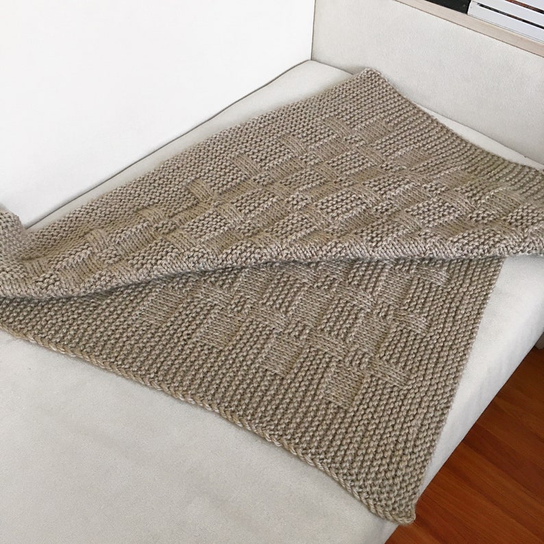 Op de afbeelding: Een beige gebreide deken met een geweven patroon. De deken is dubbelgevouwen en ligt op een witte bank.