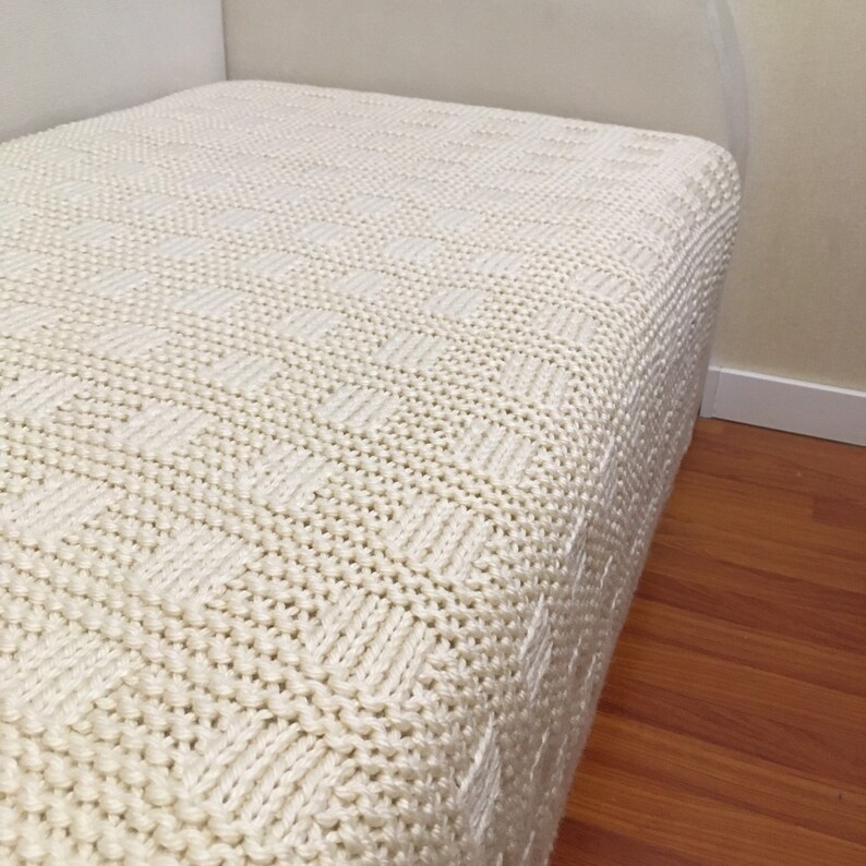 May include: A white knitted throw blanket with a textured pattern. The blanket is folded and placed on a bed.