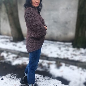 May include: A woman wearing a brown knitted sweater with a loose fit and long sleeves. She is wearing blue jeans and black boots. The photo is taken outdoors in a snowy setting.