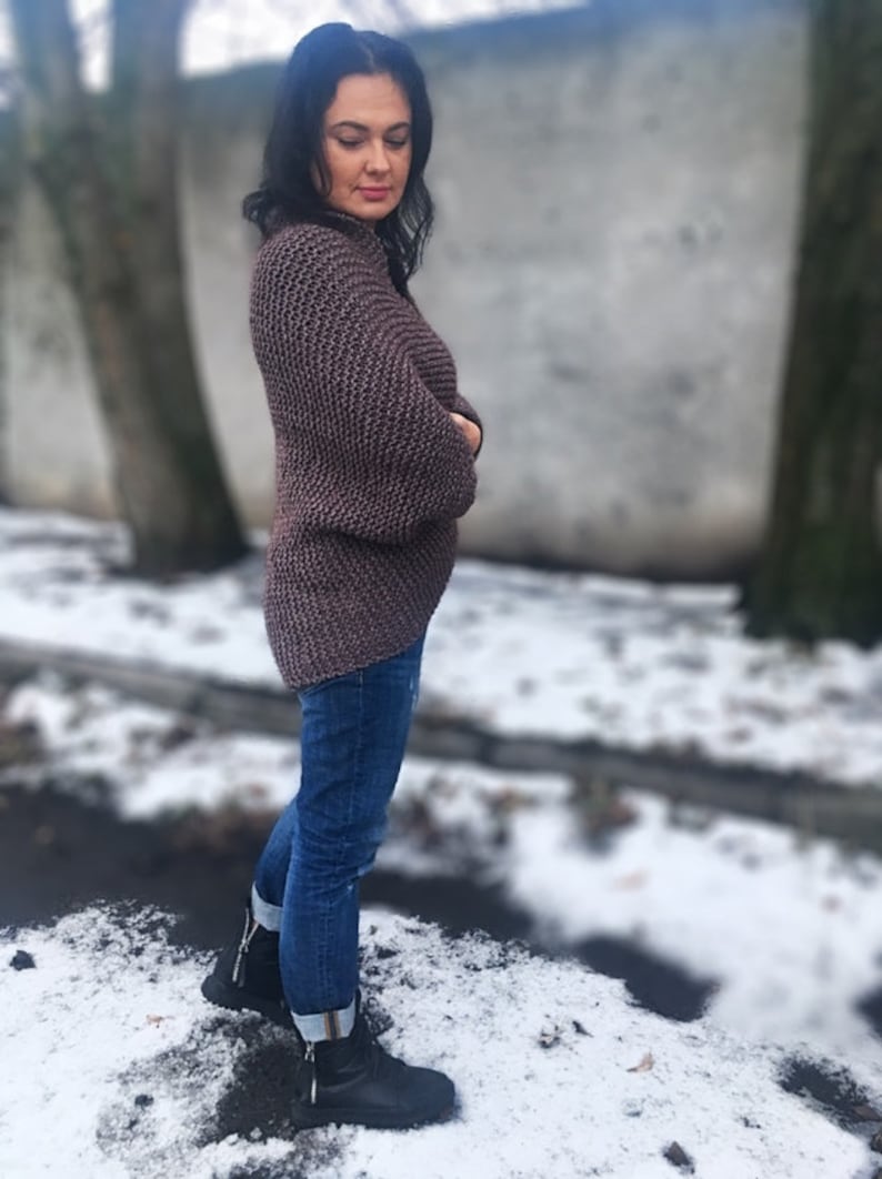 May include: A woman wearing a brown knitted sweater with a loose fit and long sleeves. She is wearing blue jeans and black boots. The photo is taken outdoors in a snowy setting.