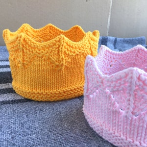 Peut inclure: Deux couronnes en tricot, l'une jaune et l'autre rose, sont présentées sur un fond en tissu gris. Les couronnes sont réalisées avec un motif de point simple et ont un sommet pointu.