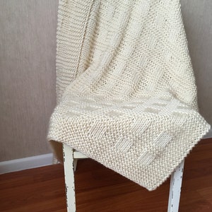 May include: A white knitted throw blanket draped over a white wooden chair. The blanket has a simple, textured pattern.