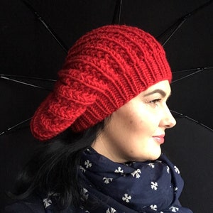 Puede incluir: Un gorro de punto rojo con un patrón de trenza. El gorro lo lleva una persona con el pelo oscuro y una bufanda azul.