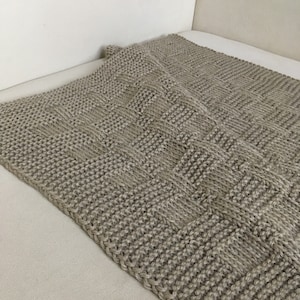 Op de afbeelding: Een beige gebreide deken met een geweven patroon. De deken is gemaakt van een zacht, getextureerd garen en heeft een subtiele glans.
