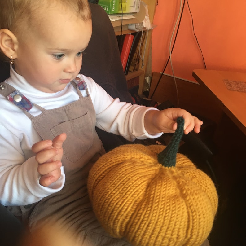 May include: A young child in a white shirt and brown overalls is holding a knitted pumpkin with a green stem. The pumpkin is yellow and has a textured surface.