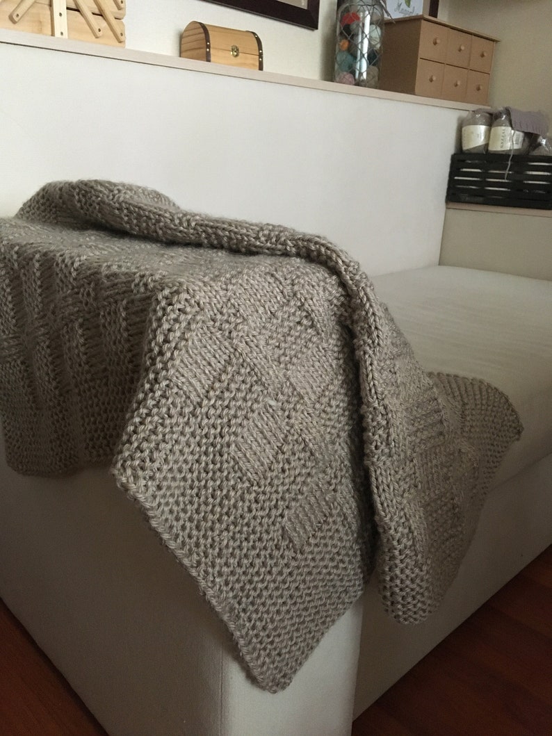 Op de afbeelding: Een beige gebreide deken met een textuurpatroon, gedrapeerd over een witte bank.