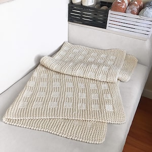 May include: A cream-colored knitted throw blanket with a textured pattern. The blanket is folded and lying on a white couch.