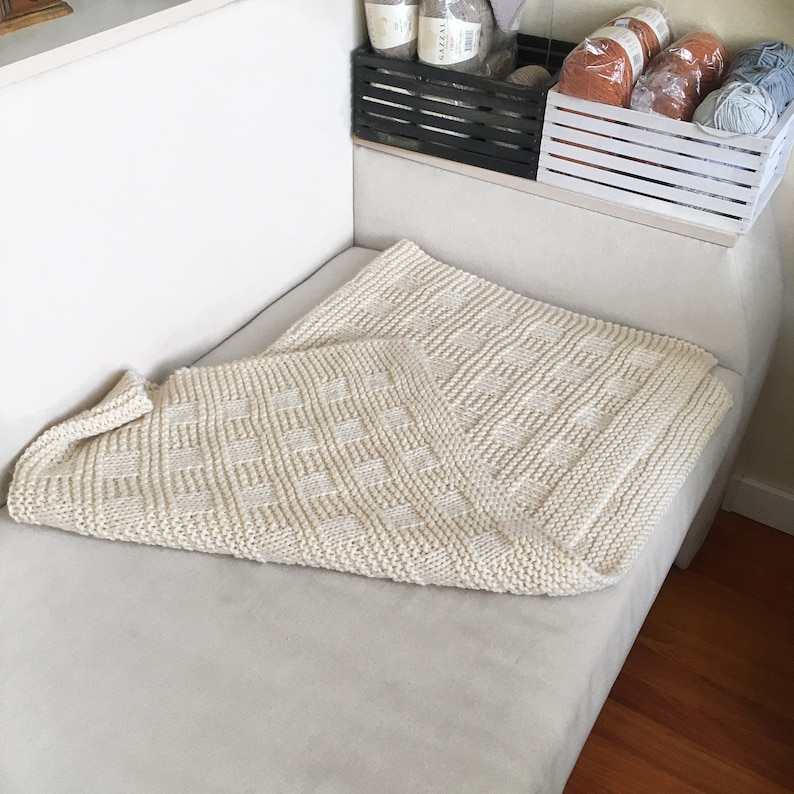 May include: A white knitted blanket with a checkered pattern, folded and placed on a white couch.