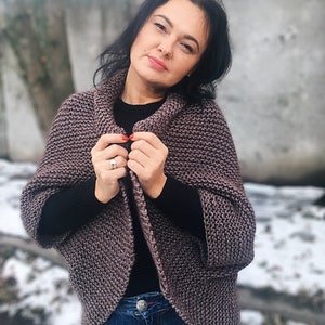 May include: A woman wearing a mauve knitted cardigan with a black shirt underneath and blue jeans. The cardigan has a cowl neck and long sleeves.