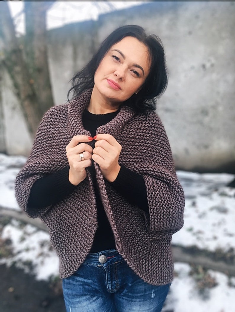 May include: A woman wearing a mauve knitted cardigan with a black shirt underneath and blue jeans. The cardigan has a cowl neck and long sleeves.