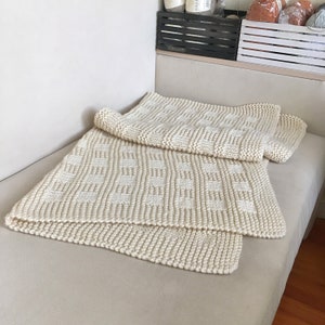 May include: A cream-colored knitted throw blanket with a geometric pattern. The blanket is folded in half and placed on a light beige couch.