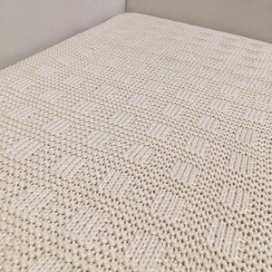May include: A close-up of a cream-colored knitted throw blanket with a textured pattern. The blanket is made of soft, cozy yarn and has a subtle sheen.
