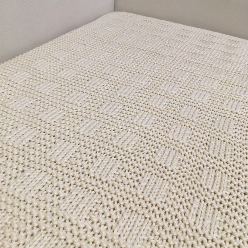May include: A close-up of a cream-colored knitted throw blanket with a textured pattern. The blanket is made of soft, cozy yarn and has a subtle sheen.