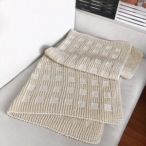 May include: A cream-colored knitted blanket with a checkered pattern. The blanket is folded and placed on a white couch.