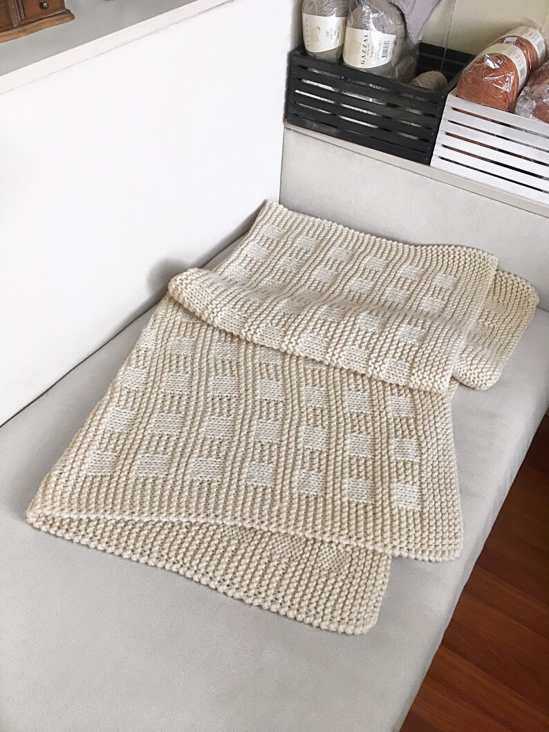 May include: A cream-colored knitted blanket with a checkered pattern. The blanket is folded and placed on a white couch.