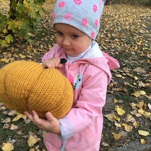 May include: A young child wearing a pink jacket and a grey and pink hat holds a knitted pumpkin in their hands. The child is standing in a pile of fallen autumn leaves.