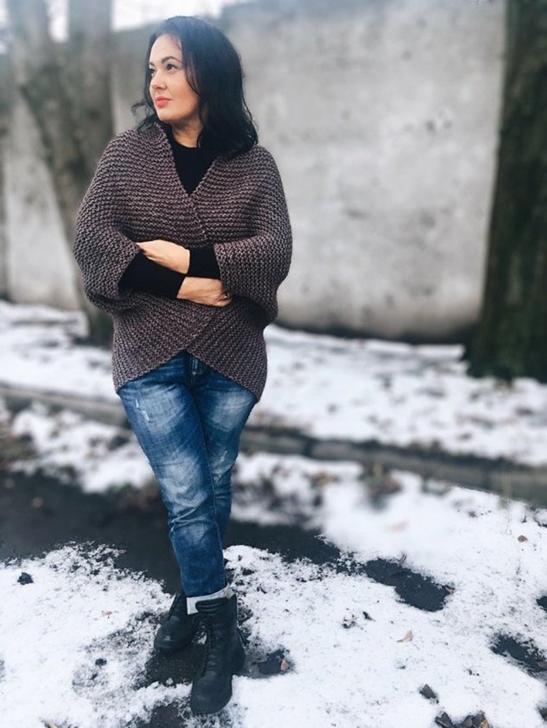 May include: A woman wearing a brown knitted cardigan with a black mock neck shirt and blue jeans. She is standing in a snowy outdoor setting.