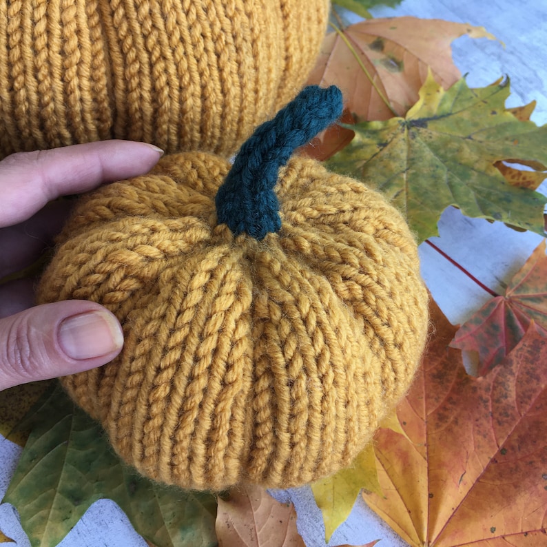 May include: Two knitted pumpkins, one larger than the other, with a green stem. The pumpkins are a deep yellow color and are sitting on a bed of fall leaves.