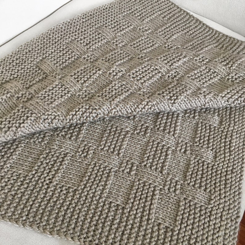 Op de afbeelding: Een beige gebreide deken met een geweven patroon. De deken is dubbelgevouwen en de textuur van de brei is zichtbaar.