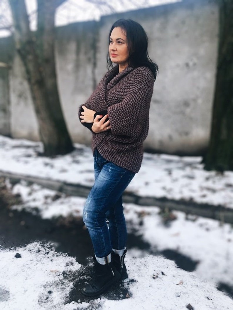 May include: A woman wearing a brown knitted cardigan with a wrap style and blue jeans. She is standing in a snowy outdoor setting.
