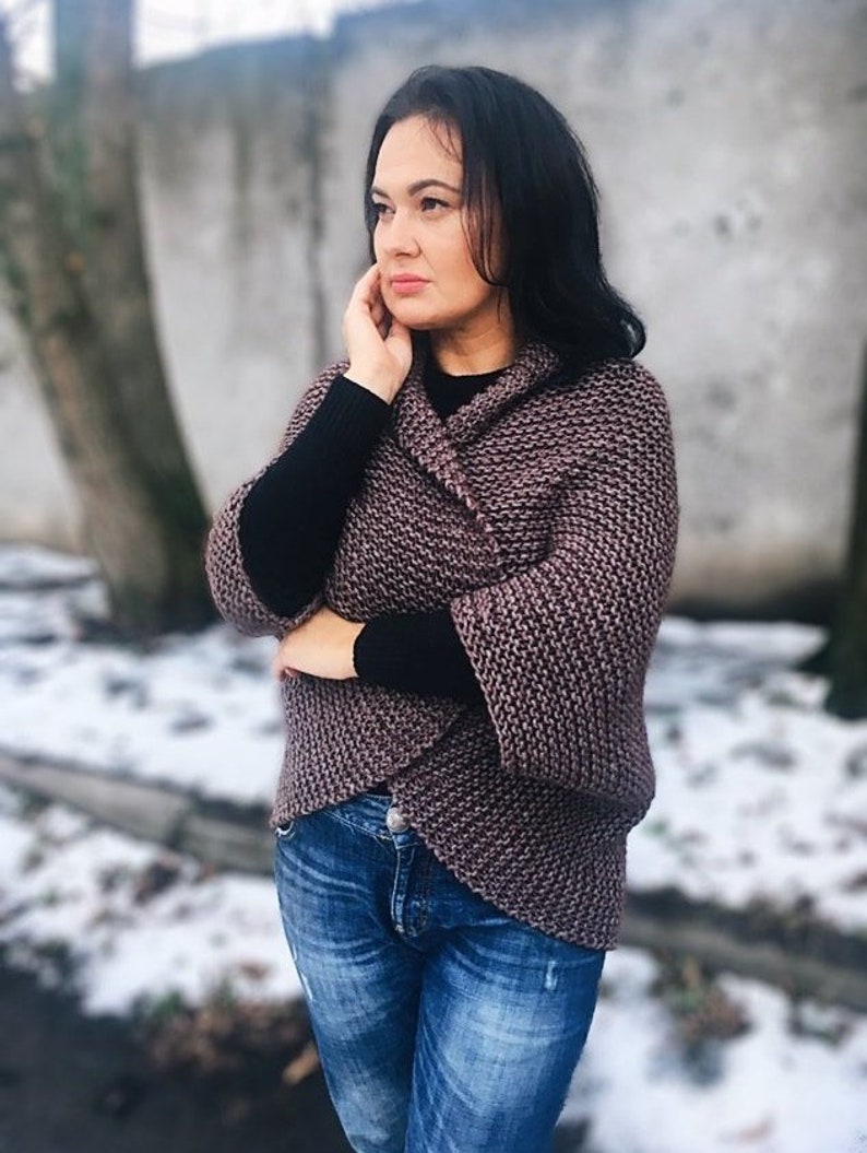 May include: A woman wearing a brown knitted cardigan with a black long-sleeved shirt underneath and blue jeans. She is standing in front of a white wall with a tree in the background.