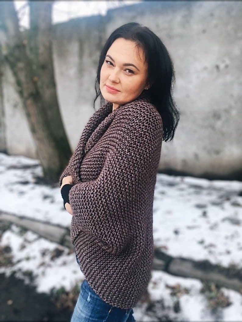 May include: A woman wearing a brown knitted cardigan with long sleeves and a relaxed fit. She is standing in front of a snowy background.